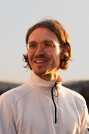 Porträt von Matthias Maurer. Er schaut lächelnd nach links. Er trägt eine Brille und einen weissen Pullover mit Reissverschluss. Seine mittellangen braunen Haare sind hinter die Ohren gekämmt.