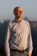 Porträt von Eric Lopsiger. Er schaut lächelnd nach rechts. Er trägt ein hellblaues Hemd mit dme obersten Knopf geöffnet.