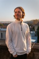Porträt von Matthias Maurer. Er schaut lächelnd nach links. Er trägt eine Brille und einen weissen Pullover mit Reissverschluss. Seine mittellangen braunen Haare sind hinter die Ohren gekämmt.