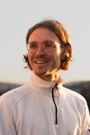 Porträt von Matthias Maurer. Er schaut lächelnd nach links. Er trägt eine Brille und einen weissen Pullover mit Reissverschluss. Seine mittellangen braunen Haare sind hinter die Ohren gekämmt.